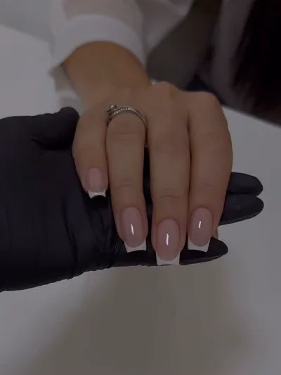 A finished square french manicure, held by the technician's gloved hand.